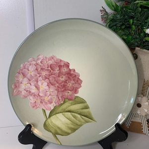 May include: A round, light green plate with a pink hydrangea flower and green leaf design. The plate has a light blue rim and is displayed on a black stand. The floral design adds a touch of nature to the tableware.