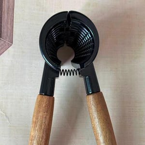 Peut inclure: Un casse-noix noir avec des poignées en bois et un ressort argenté. Le casse-noix a une tête circulaire avec un intérieur strié. Les poignées sont brun clair et inclinées vers l'extérieur.