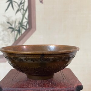 May include: A bronze-colored bowl with intricate floral and geometric patterns. The bowl sits on a small, dark red wooden stand. The background features a framed painting of bamboo stalks.
