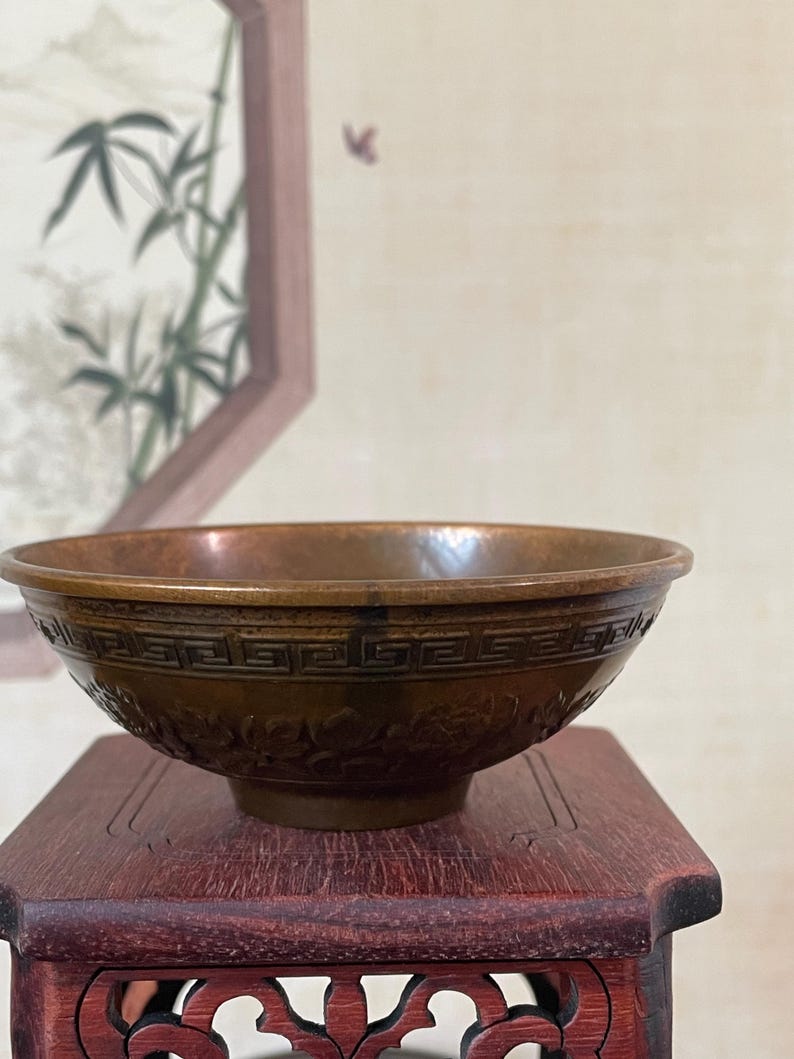 May include: A bronze-colored bowl with intricate floral and geometric patterns. The bowl sits on a small, dark red wooden stand. The background features a framed painting of bamboo stalks.