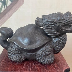 May include: A dark gray sculpture of a mythical creature with a turtle-like body and a dragon-like head. The sculpture is placed on a dark red wooden base. The details of the sculpture are finely carved, showcasing the texture of the scales and the creature's features.