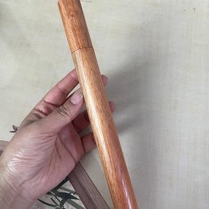 Può includere: Un contenitore cilindrico in legno con coperchio. Il contenitore è realizzato in legno marrone rossiccio con venature visibili. Il coperchio si adatta perfettamente sopra. Il contenitore è tenuto in mano, suggerendo la sua natura portatile. Il legno ha una finitura lucida.