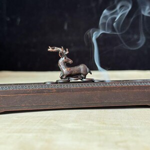 Puede incluir: Un quemador de incienso de bronce con una base rectangular decorada y una pequeña figura de ciervo. El humo se eleva del quemador, lo que indica el uso de incienso. El diseño es ornamentado, adecuado para la decoración del hogar. La base mide unos 20 cm de largo.