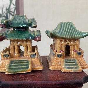 May include: Two ceramic pagoda figurines. The structures feature a multi-tiered green roof, tan walls, and a small staircase. The pagoda on the left has a more ornate roof design. The figurines are on a dark wooden surface.