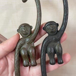 May include: Two bronze-colored monkey-shaped hooks. The monkeys are depicted hanging with their tails curved upwards. The hooks have a detailed, textured finish, and the monkeys have smiling faces. The hooks are likely for decorative or functional use.