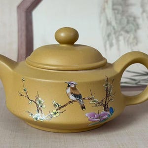 May include: Mustard-yellow ceramic teapot with a lid and handle. The teapot is decorated with a painted bird perched on a branch with flowers. The teapot has a capacity of approximately 1.2 litres.