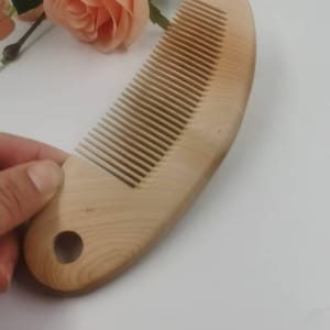 Puede incluir: Un peine de madera de color claro con forma curva y un orificio circular en la base del mango. El peine presenta dientes finos y espaciados uniformemente, con un acabado suave que muestra la veta natural de la madera.