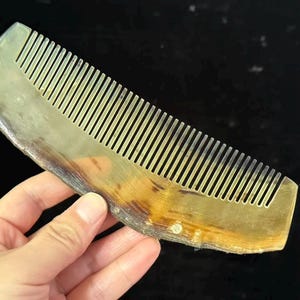 Puede incluir: Un peine de cuerno con una forma natural y orgánica. El peine tiene un color marrón claro y beige con una serie de dientes finos y paralelos. El peine se sostiene sobre un fondo oscuro.
