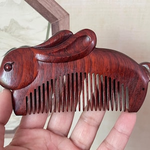 Puede incluir: Un peine de madera tallado en forma de conejo. El peine es de madera marrón rojiza y presenta un diseño detallado de la cabeza, las orejas y el cuerpo del conejo. Los dientes del peine están espaciados uniformemente. Un accesorio único para el cuidado del cabello, de unos 15 cm.