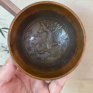 May include: A small, round, bronze-colored bowl with an embossed design of two koi fish and Chinese characters. The bowl has a smooth, polished exterior and a detailed interior with the fish and floral elements. The bowl is held in a hand.