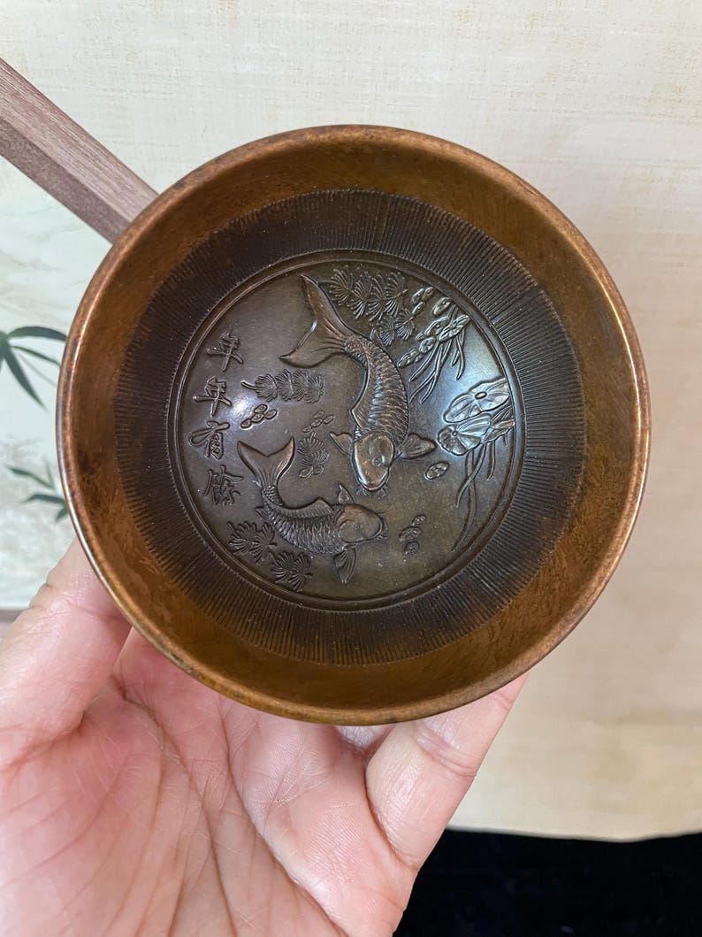 May include: A small, round, bronze-colored bowl with an embossed design of two koi fish and Chinese characters. The bowl has a smooth, polished exterior and a detailed interior with the fish and floral elements. The bowl is held in a hand.