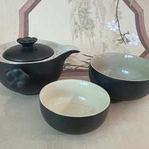 Peut inclure: Service à thé en céramique noire comprenant une théière et deux bols assortis. La théière a un couvercle noir et une poignée en forme de fleur. Les bols ont un extérieur noir et un intérieur blanc. L'ensemble est conçu pour les cérémonies du thé.
