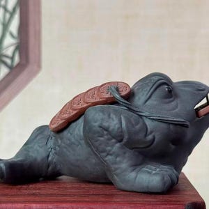 Könnte beinhalten: Dunkelgraue Keramik-Froschfigur mit einer Münze im Maul und einem dekorativen Stück auf dem Rücken. Die liegende Froschfigur steht auf einer roten Holzoberfläche. Die Figur hat detaillierte Merkmale und eine glatte Oberfläche.