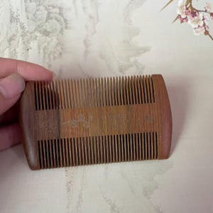 May include: A rectangular wooden comb featuring fine teeth on both the top and bottom edges. The center of the comb displays a delicate laser-engraved design of butterflies and floral vines across the natural brown wood grain surface.