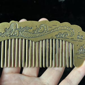 May include: A bronze-colored comb featuring a mermaid design. The comb has a decorative top with an engraved mermaid, waves, and flowing lines. The comb's teeth are vertical and evenly spaced.