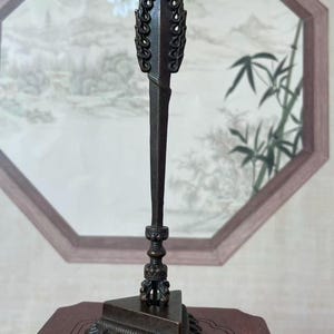 Peut inclure: Un poignard rituel phurba de couleur bronze foncé doté d'un sommet orné en forme de flamme, d'une tige centrale lisse et effilée, et d'un manche décoratif aux détails circulaires complexes, monté sur une base triangulaire solide.