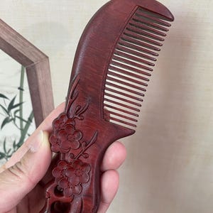 Puede incluir: Un peine de madera marrón rojizo con un mango curvo y dientes finos. El mango está intrincadamente tallado con diseños florales. El peine se sostiene en una mano, mostrando la artesanía detallada y la veta natural de la madera.
