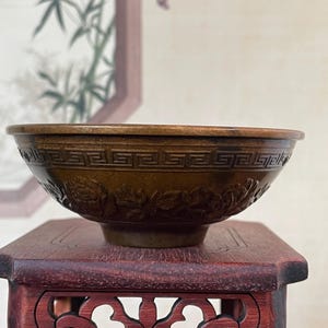 May include: A brown, antique-style bowl with intricate carved designs. The bowl features a geometric border and floral patterns. It sits on a small, dark red wooden stand with decorative cutouts. The background includes a framed painting.