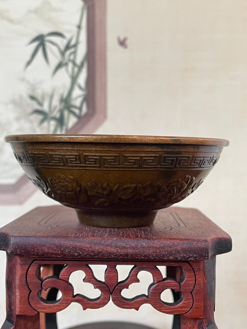 May include: A brown, antique-style bowl with intricate carved designs. The bowl features a geometric border and floral patterns. It sits on a small, dark red wooden stand with decorative cutouts. The background includes a framed painting.