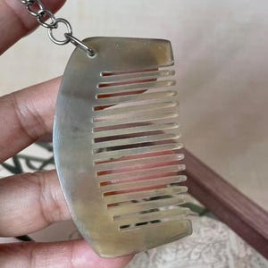 Pode incluir: Um pequeno pente porta-chaves em chifre, semitranslúcido e de cor castanho-claro, com um dorso curvo e liso e uma série de dentes espaçados uniformemente. O pente está preso a uma corrente e argola de metal prateado para uso como acessório.