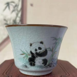 May include: A small, light blue ceramic teacup with a panda bear illustration. The panda is black and white, holding bamboo leaves. The cup has a crackle glaze and a brown rim. The cup is sitting on a wooden surface.