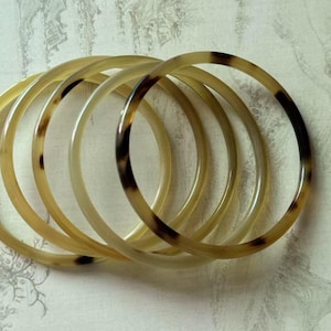 Könnte beinhalten: Eine Sammlung von sechs durchscheinenden, kreisförmigen Armreifen. Die Armreifen haben einen hellgelben Farbton mit braunen und schwarzen Marmorierungen. Der Schmuck ist übereinander gestapelt, wodurch ein geschichteter Effekt entsteht. Der Hintergrund ist eine helle, strukturierte Oberfläche.