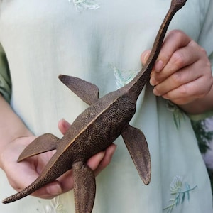 May include: Bronze sculpture of a plesiosaur, a prehistoric marine reptile. The sculpture has a long neck, flippers, and a textured surface. The color is a dark bronze with a mottled appearance. The sculpture is held in someone's hands.