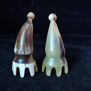 May include: Two decorative, abstract sculptures. One is brown and white, the other is pale green and brown. Both have a rounded top and a base with three curved prongs. The sculptures are set against a dark background.