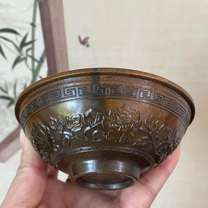 May include: Bronze-colored bowl with intricate floral and geometric patterns. The bowl features a detailed relief of flowers around the exterior and a repeating geometric design near the rim. The bowl is held in a hand, showcasing its size and craftsmanship.
