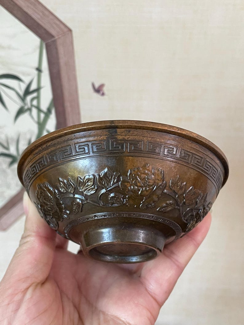 May include: Bronze-colored bowl with intricate floral and geometric patterns. The bowl features a detailed relief of flowers around the exterior and a repeating geometric design near the rim. The bowl is held in a hand, showcasing its size and craftsmanship.