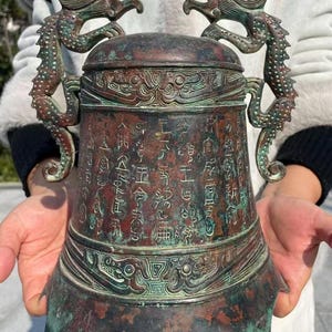 May include: An antique bronze bell with intricate carvings and inscriptions. Two dragon figures adorn the top, and the bell has a handle. The bell's surface shows a patina of green and brown hues, suggesting age and use.