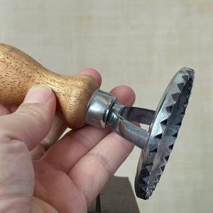Op de afbeelding: Een ravioli-stempel voor in de keuken met een gladde, lichtbruine houten handgreep bevestigd aan een ronde, zilverkleurige metalen voet. De metalen voet heeft een opvallende, scherpe, gekartelde of gegroefde rand voor het snijden van deeg.