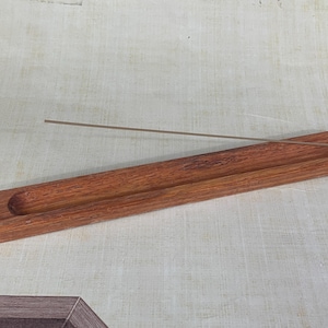 Könnte beinhalten: Ein rechteckiger Räucherstäbchenhalter aus Holz mit einer Nut zur Aufnahme von Räucherstäbchen. Der Halter ist aus rotbraunem Holz gefertigt und hat ein schlichtes, minimalistisches Design. Ein Räucherstäbchen ist in dem Halter platziert.