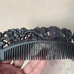 Puede incluir: Un peine de madera negra tallado intrincadamente. El peine presenta un diseño floral y de nubes a lo largo del borde superior. Los dientes están espaciados uniformemente. El peine se sostiene en una mano.