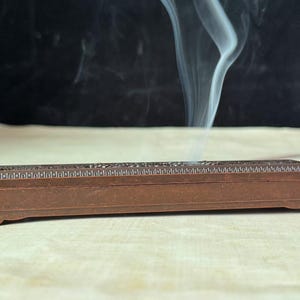 Puede incluir: Un quemador de incienso rectangular marrón con intrincados detalles tallados. Volutas de humo blanco se elevan del quemador sobre un fondo oscuro, lo que sugiere el uso de incienso. El quemador está sobre una superficie clara.