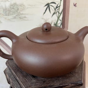 Peut inclure: Une théière en céramique marron avec un corps arrondi, une anse incurvée et un bec verseur. Le couvercle a un petit bouton. La théière est exposée sur un support en bois foncé. L'arrière-plan comprend une peinture encadrée.