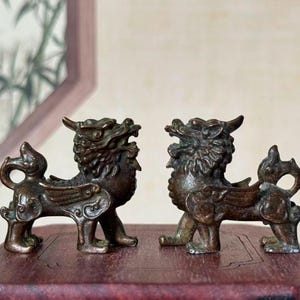 May include: Two bronze-colored mythical creatures, possibly Foo Dogs or similar guardians, are displayed on a wooden surface. Each figurine has a detailed, textured design with wings, horns, and an open mouth, suggesting a protective or decorative function.