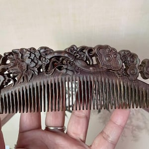 Puede incluir: Un peine de madera marrón oscuro con intrincadas tallas de flores y nubes. El mango del peine presenta diseños florales y de nubes detallados, y los dientes están finamente espaciados. Un accesorio decorativo para el cabello.