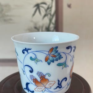 Puede incluir: Taza de cerámica blanca con un diseño floral en azul, naranja y verde. La taza presenta una forma curva y un patrón detallado de flores y vides. El fondo está borroso.