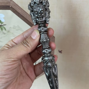 May include: An intricately carved silver dagger. The handle features a multi-faced deity design, a crown, and skull motifs. The blade is pointed. The dagger is held in a hand. The overall design suggests a ceremonial or decorative purpose.