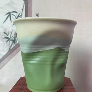 Pode incluir: Um vaso de cerâmica artesanal com um design em camadas. A parte superior é creme esbranquiçado, passando para um verde suave e terminando com uma base verde vibrante. O vaso tem uma forma ligeiramente texturizada e orgânica.