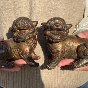 May include: Two antique bronze-colored lion statues. Each statue is in a seated position with a detailed mane, open mouth, and a reddish-brown hue. The statues are likely decorative items.