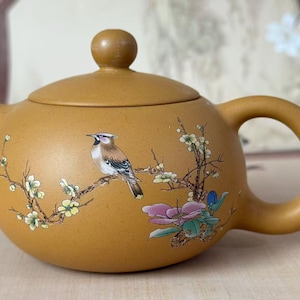 Puede incluir: Una tetera de cerámica de color mostaza con tapa y asa. La tetera está decorada con un pájaro pintado posado en una rama floreciente con flores rosas y blancas. La tetera tiene forma redondeada.