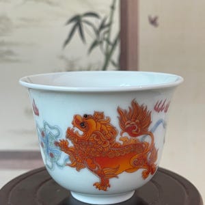 May include: White ceramic teacup with a vibrant orange and red lion design. The cup features a detailed illustration of a mythical creature, with blue and red accents. The cup is set on a dark wooden stand, showcasing the intricate artwork.