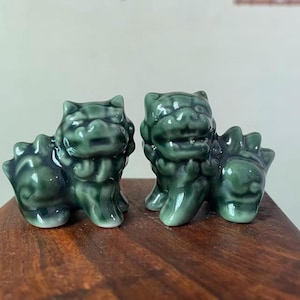 May include: Two ceramic guardian lion statues in a glossy, dark green glaze. The statues are detailed with stylized features, including a mane, paws, and a tail. They are placed on a wooden surface.