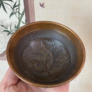May include: A small, bronze-colored bowl with an embossed design of two koi fish swimming among aquatic plants. The bowl's interior features a detailed relief, and the rim has a darker, aged appearance. The bowl is held in a hand.