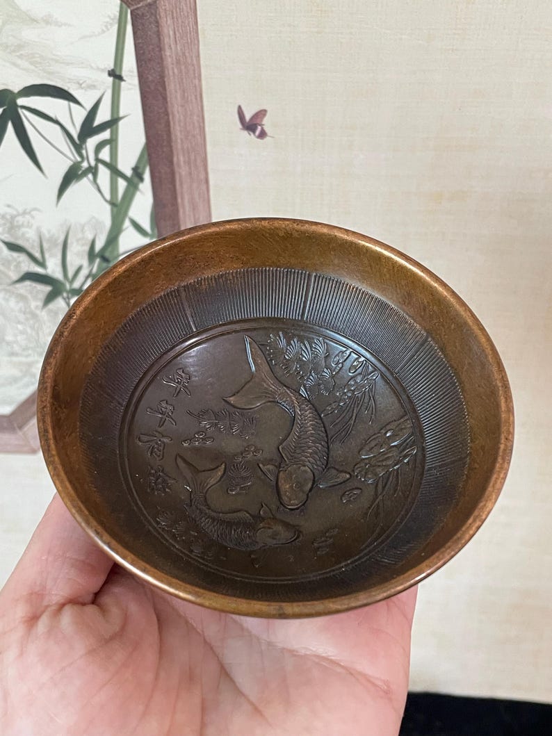 May include: A small, bronze-colored bowl with an embossed design of two koi fish swimming among aquatic plants. The bowl's interior features a detailed relief, and the rim has a darker, aged appearance. The bowl is held in a hand.