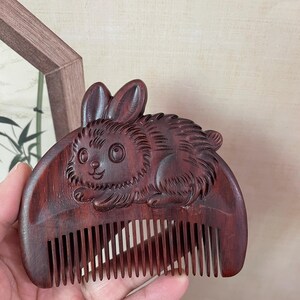 Puede incluir: Un peine de madera de color marrón rojizo oscuro con un diseño de conejo tallado. El peine tiene dientes espaciados uniformemente y una talla detallada de un conejo en la parte superior. El conejo tiene detalles.