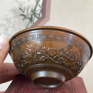 May include: A bronze-colored bowl with intricate floral and geometric patterns. The bowl features detailed carvings of flowers and leaves around the exterior, with a repeating geometric design near the rim. The bowl is displayed on a dark wooden stand.