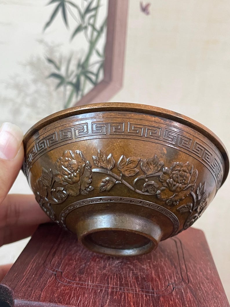 May include: A bronze-colored bowl with intricate floral and geometric patterns. The bowl features detailed carvings of flowers and leaves around the exterior, with a repeating geometric design near the rim. The bowl is displayed on a dark wooden stand.